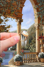 Load image into Gallery viewer, Edited background is of a painted castle balcony looking over onto a beautiful lake and waterfall scene. Lots of flowers and plants hanging off the balcony. In the forefront is a hand holding with two fingers the Isabella ring, she is a wire wrapped gold filled ring with a coin shaped freshwater pearl as her feature.
