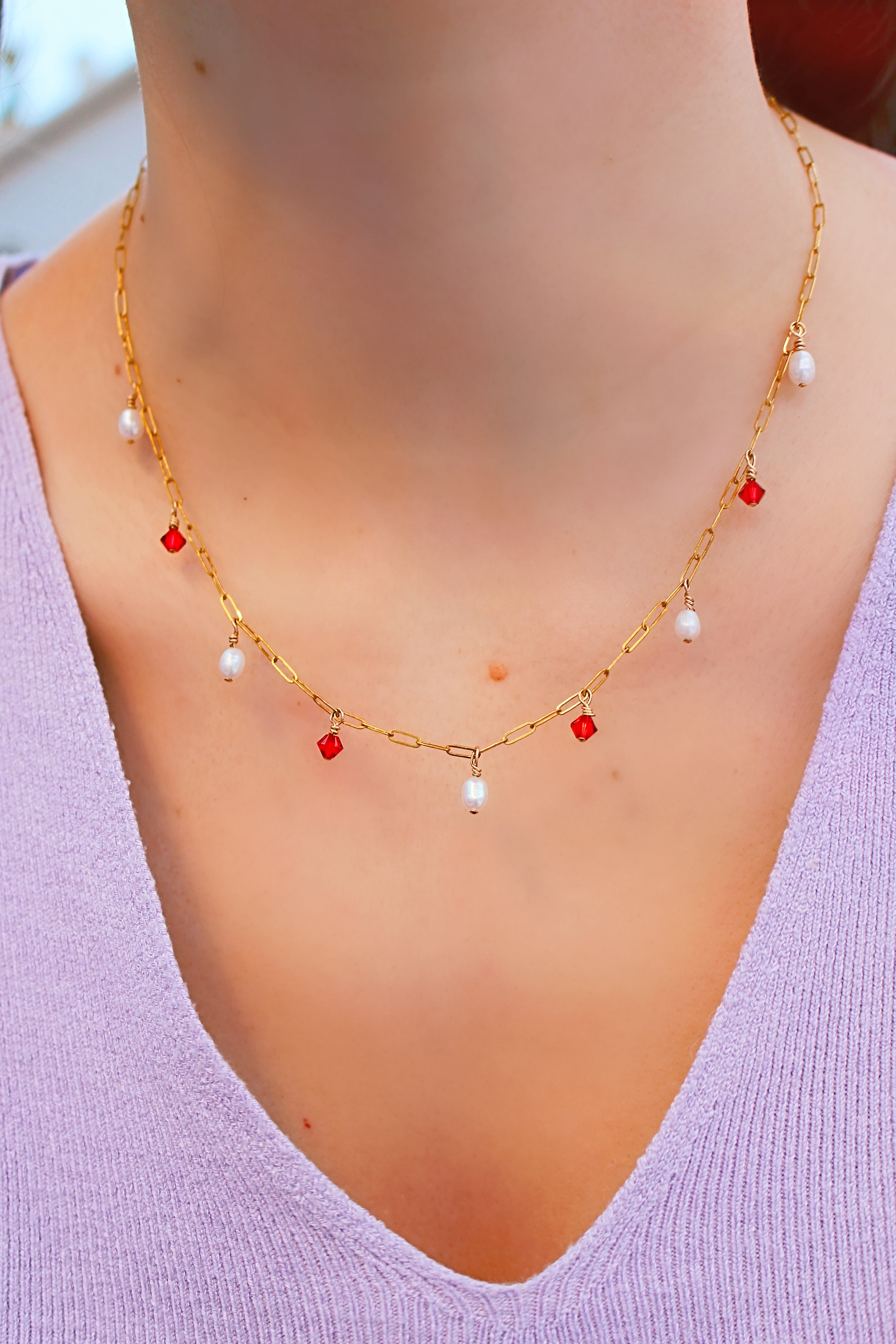 Lady wearing a lilac v-neck top wearing the Anastasia necklace, the frame is focused on the chest area to bring focus to the necklace. The necklace is a gold filled satelite chain with five pearls and four red swarovski crystals interchanging.
