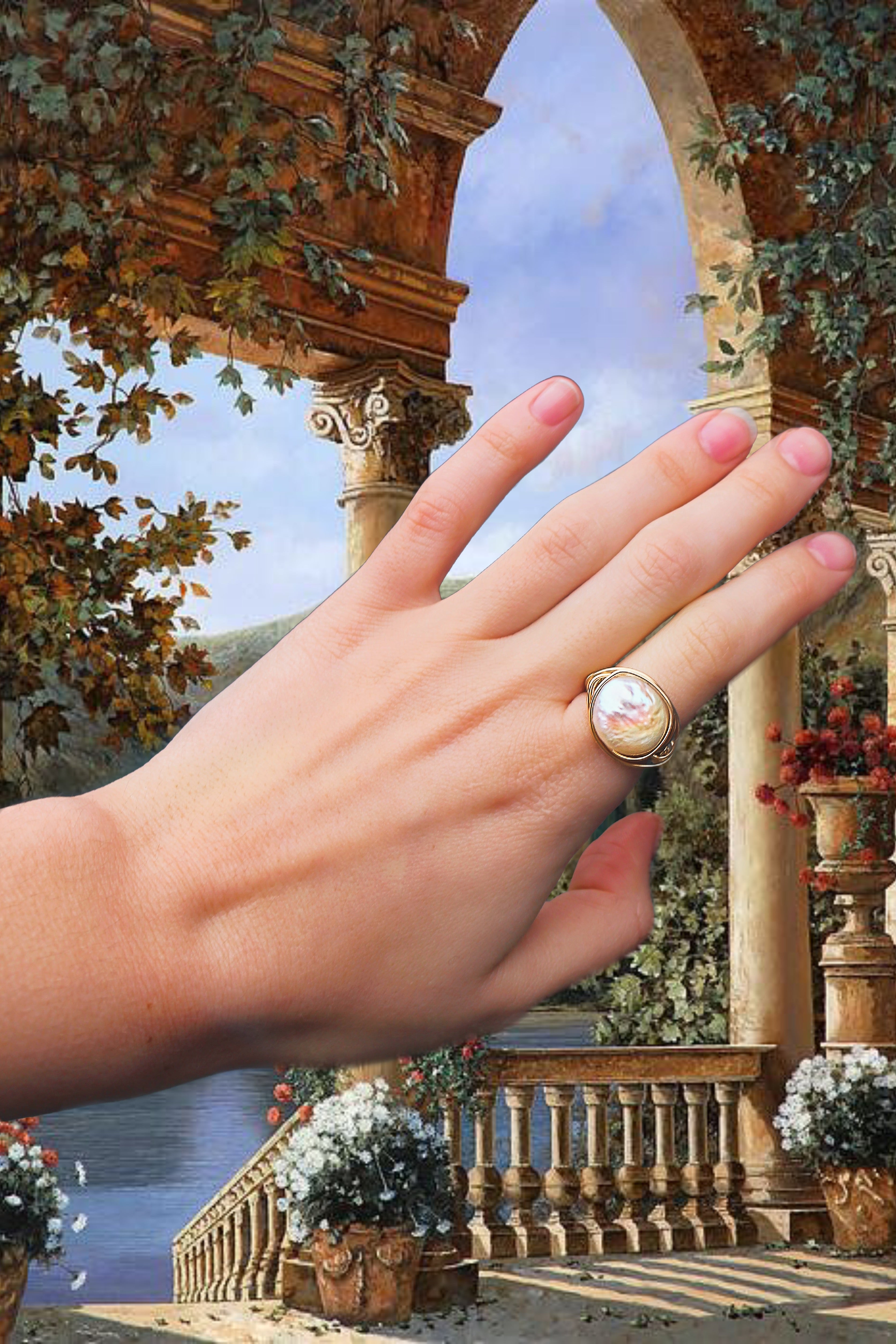 Edited background is of a painted castle balcony looking over onto a beautiful lake and waterfall scene. Lots of flowers and plants hanging off the balcony. In the forefront is a hand wearing the Isabella ring on the pointer finger, she is a wire wrapped gold filled ring with a coin shaped freshwater pearl as her feature.