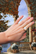 Load image into Gallery viewer, Edited background is of a painted castle balcony looking over onto a beautiful lake and waterfall scene. Lots of flowers and plants hanging off the balcony. In the forefront is a hand wearing the Isabella ring on the pointer finger, she is a wire wrapped gold filled ring with a coin shaped freshwater pearl as her feature.
